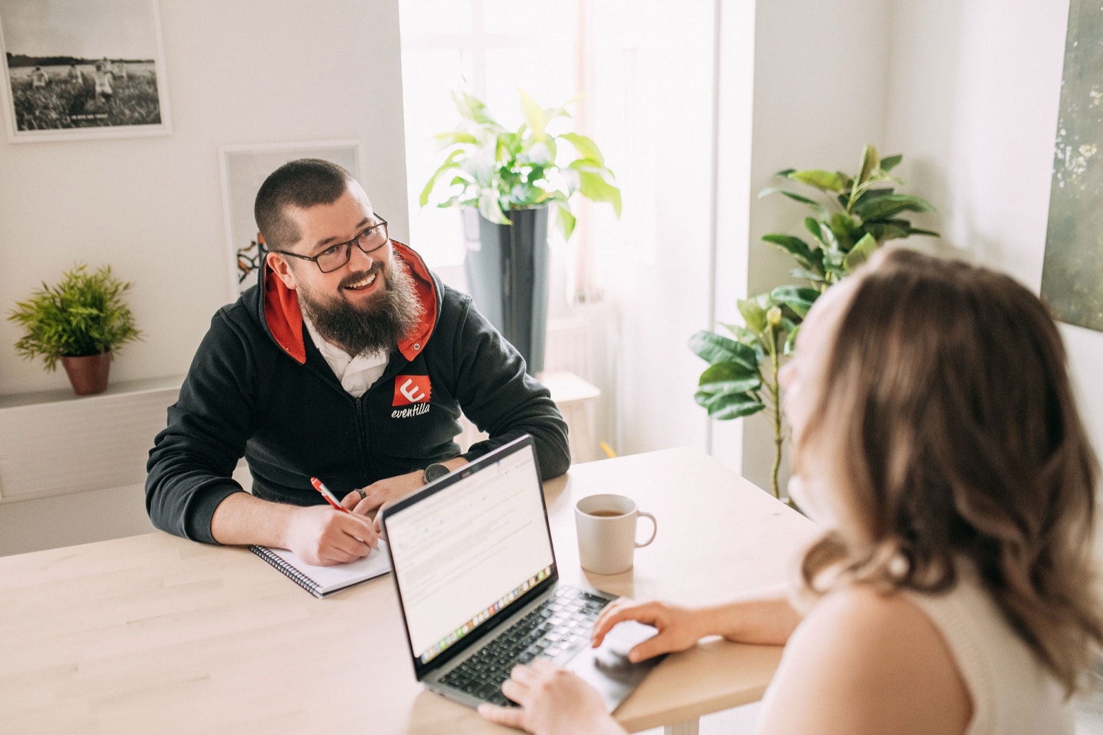 A CompetenceFlow team member talking with a visitor across a desk with a laptop and coffee in a bright office in Oulu, Finland